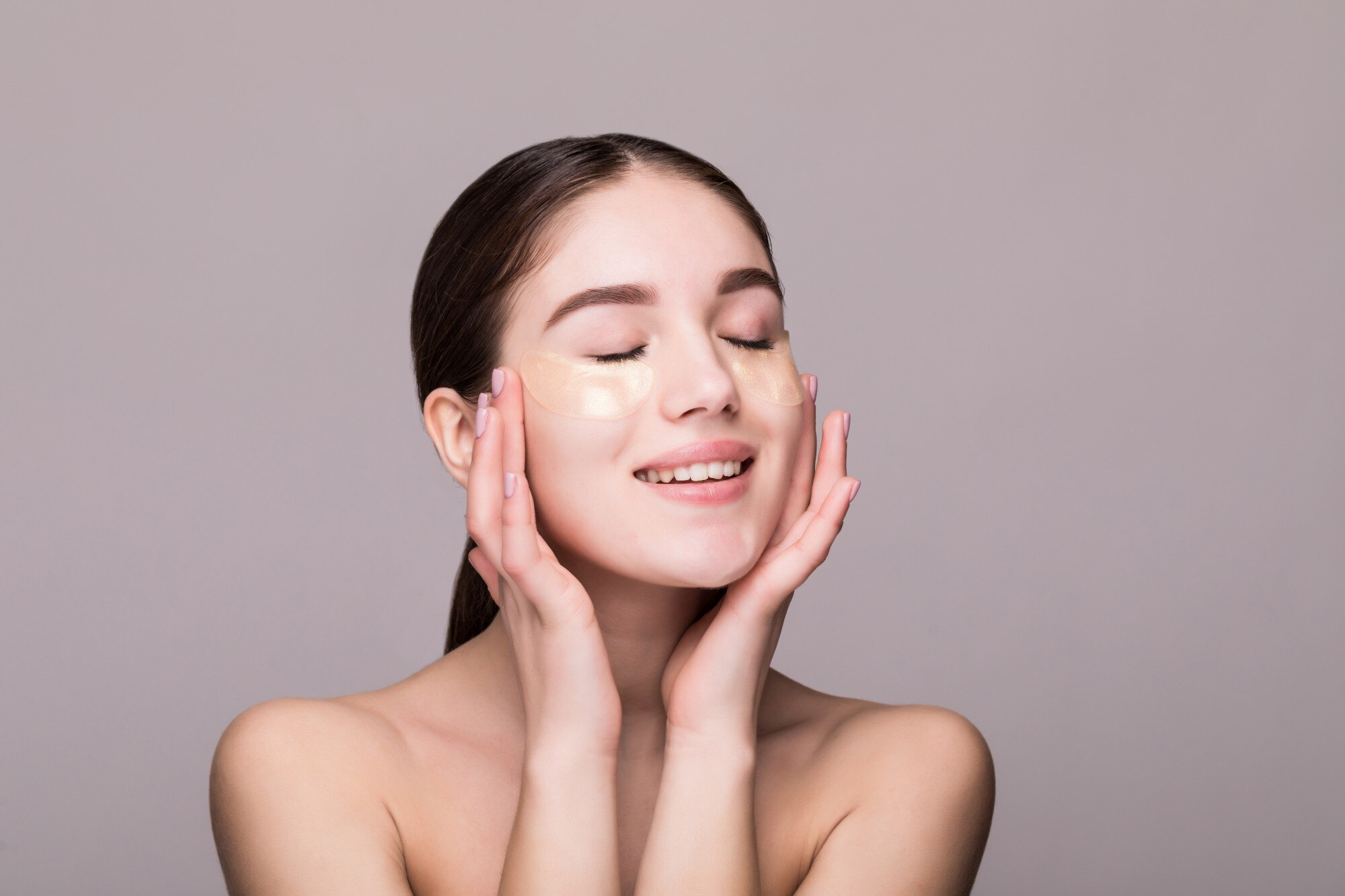 Young woman with an eye patches touching temples isolated on gray wall. Cosmetics